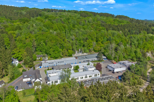 Luftaufnahme eines Produktionsstandorts mit mehreren Industriegebäuden, umgeben von dicht bewaldeten Hügeln