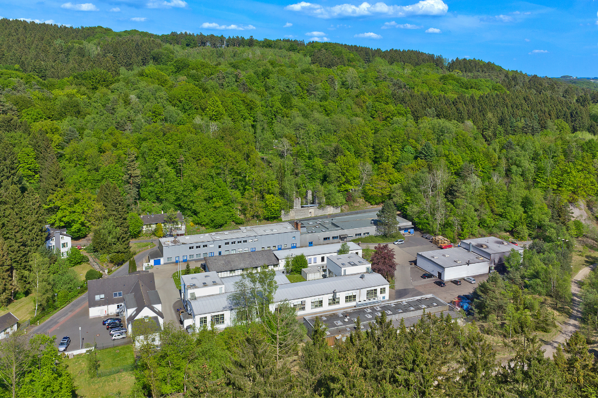 Luftaufnahme eines Produktionsstandorts mit mehreren Industriegebäuden, umgeben von dicht bewaldeten Hügeln