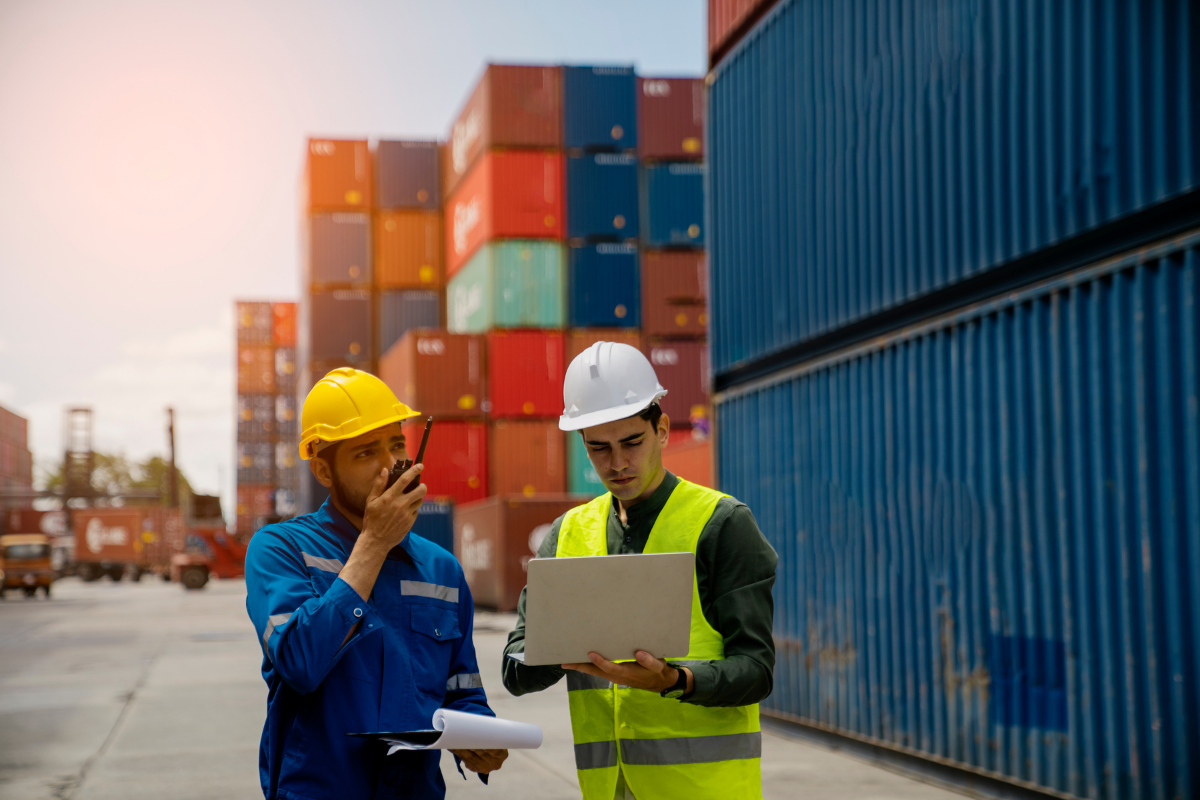 Zwei Logistikmitarbeiter mit Schutzhelmen und Warnweste arbeiten mit Laptop und Funkgerät vor gestapelten Frachtcontainern